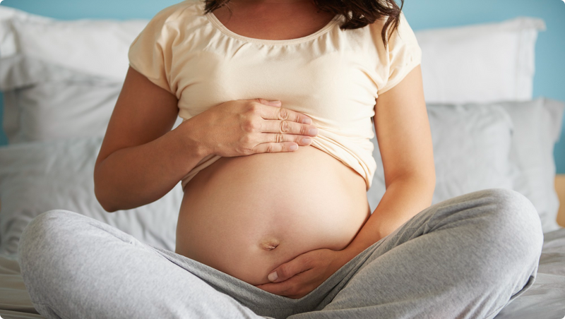 A pregnant person sitting cross-legged on a bed, wearing a light-colored shirt and gray pants, with hands gently resting on their bare belly. The background shows pillows and bedding.