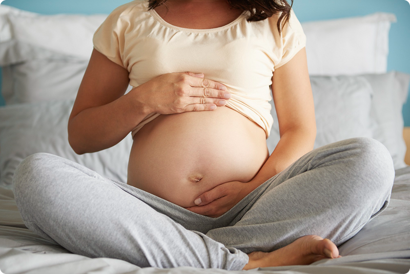 A pregnant person sitting cross-legged on a bed, gently holding their bare belly with both hands. They are wearing a light-colored shirt and gray sweatpants. The background shows pillows and a soft blue wall.