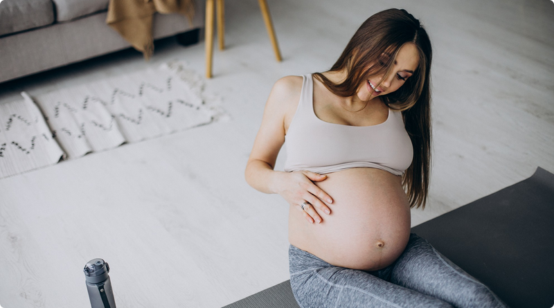 Pregnant woman sitting on a yoga mat at home, smiling and gently touching her belly, wearing a light sports bra and gray leggings.