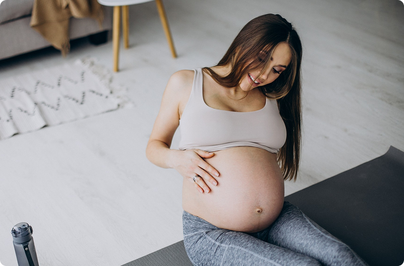 A pregnant woman in workout clothes sits on a yoga mat, smiling as she gently cradles her bare belly with both hands in a bright, cozy room.