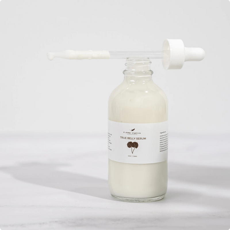 A clear glass bottle filled with white serum labeled True Belly Serum stands on a light surface. The dropper lid rests on top, with some serum visible on the dropper wand.