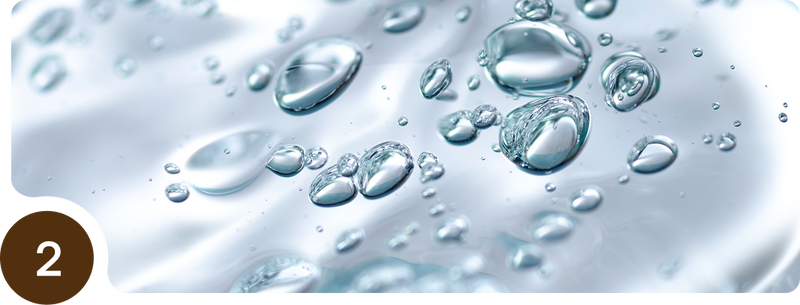 Close-up of clear water bubbles suspended in liquid against a soft, light background. In the bottom left corner, there is a brown circle with the number 2 inside it.