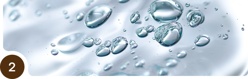 Close-up image of clear water droplets suspended in a transparent liquid, reflecting light and creating a shiny, smooth texture. A brown circle with the number 2 appears in the lower left corner.
