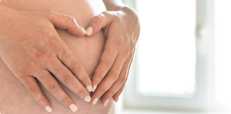 Close-up of two hands forming a heart shape on a pregnant belly, with a softly lit window in the background.