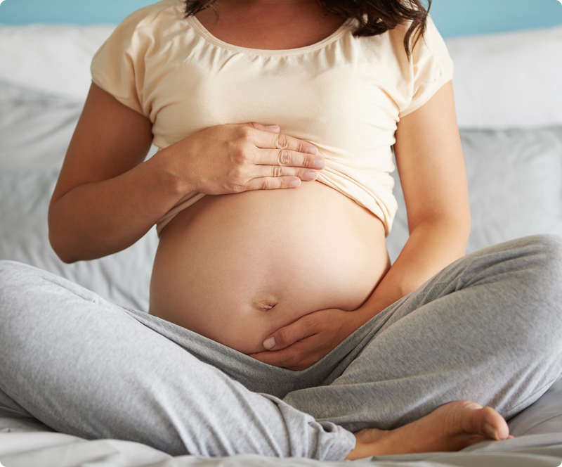 A pregnant woman sits cross-legged on a bed, gently cradling her bare belly with both hands. She wears a light beige top and gray sweatpants, and the background features neutral bedding and pillows.