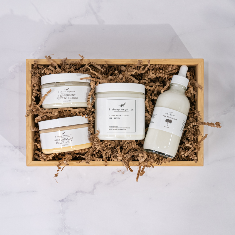 A top-down view of an 8 Sheep Organics  set in a wooden box with brown paper filler. The set includes four products: Sleepy Body Lotion, True Belly Serum, Peppermint Foot & Leg Rub, and Helichrysum Belly Balm.