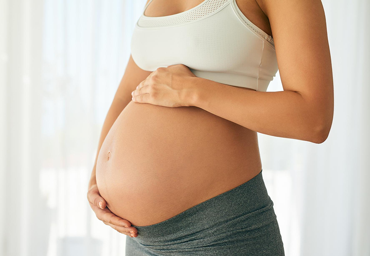 A pregnant person wearing a sports bra and leggings gently cradles their bare belly with both hands. The background is softly lit with natural light.