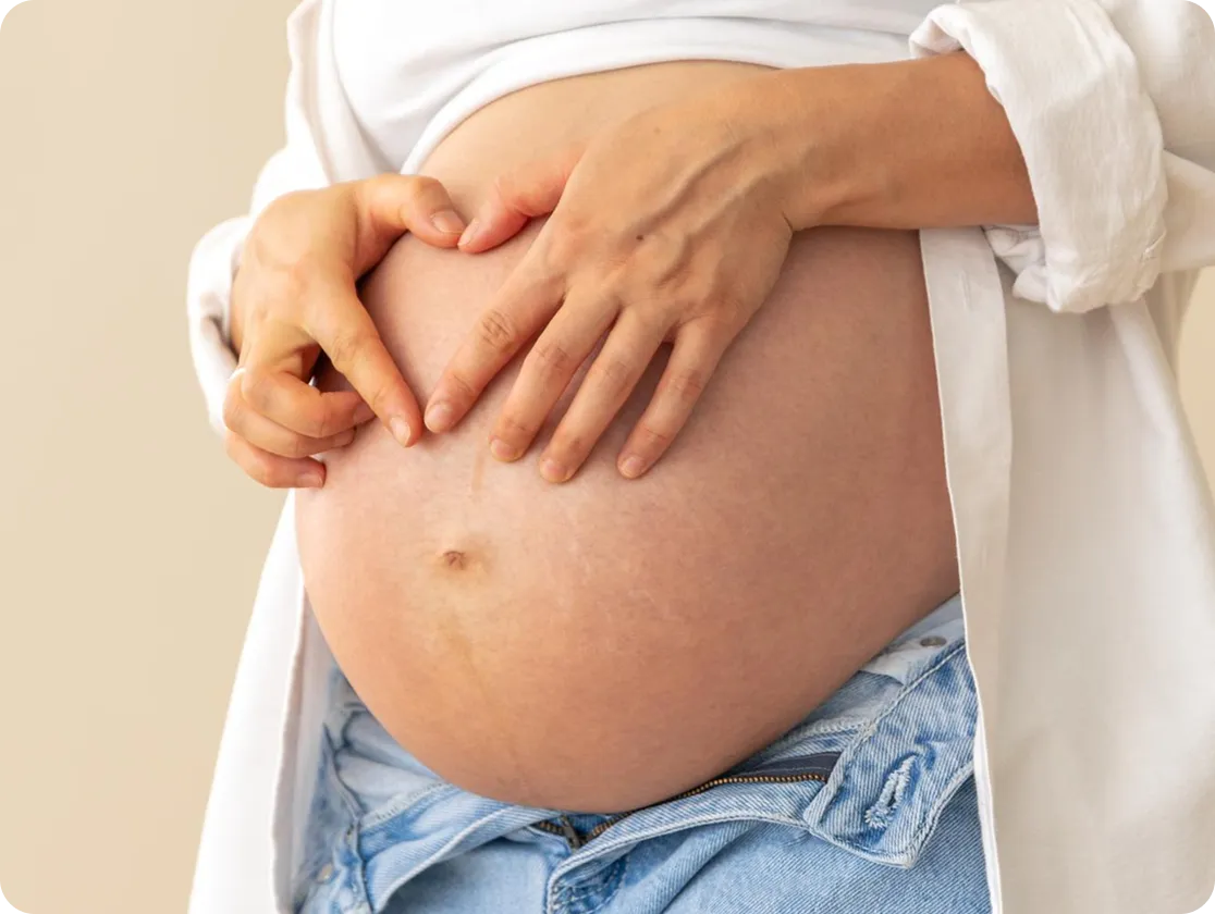 A close-up of a pregnant person’s bare belly with their hands gently forming a heart shape, wearing an open white shirt and blue jeans.