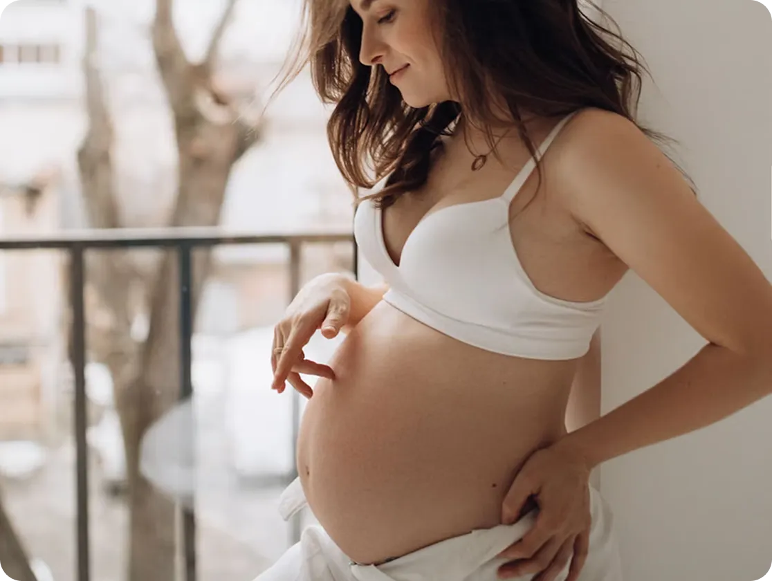 A pregnant woman wearing a white bra and shirt open at the waist stands by a window, gently touching her belly and smiling softly. Natural light fills the room, creating a calm atmosphere.