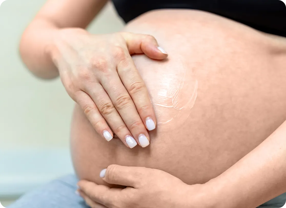 A close-up of a pregnant person applying lotion to their bare belly with one hand while supporting it with the other hand. The focus is on the belly and hands.