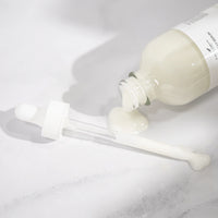 A glass dropper and bottle with white lotion are lying on a light surface; some creamy liquid has spilled out of the bottle onto the surface near the dropper.