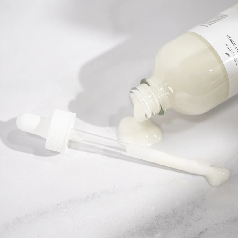 A glass dropper and bottle with white lotion are lying on a light surface; some creamy liquid has spilled out of the bottle onto the surface near the dropper.